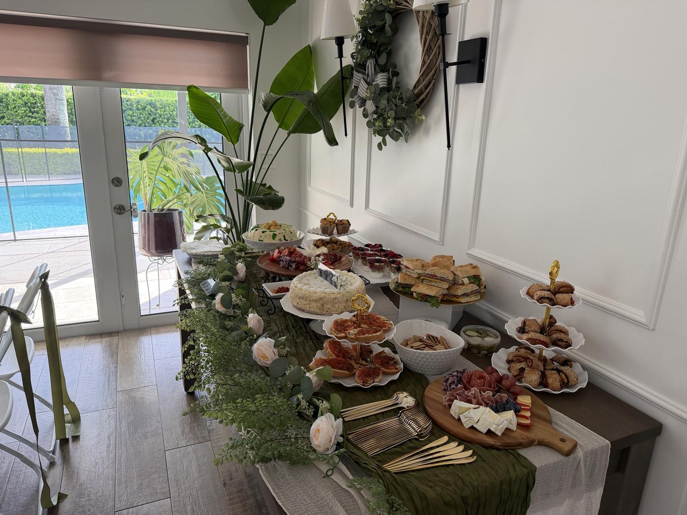 Aura Gourmet event table by the pool with charcuterie, sandwiches, pastries, cake and a greenery runner
